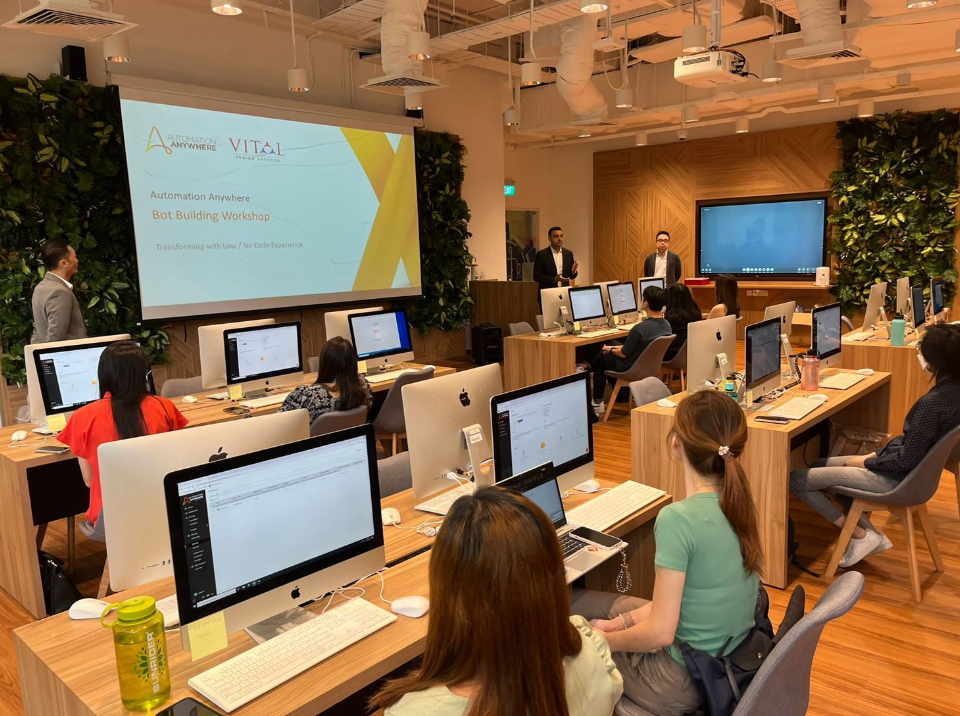 Participants sit at iMac computers during an Automation Anywhere Bot Building Workshop with two presenters at the front.